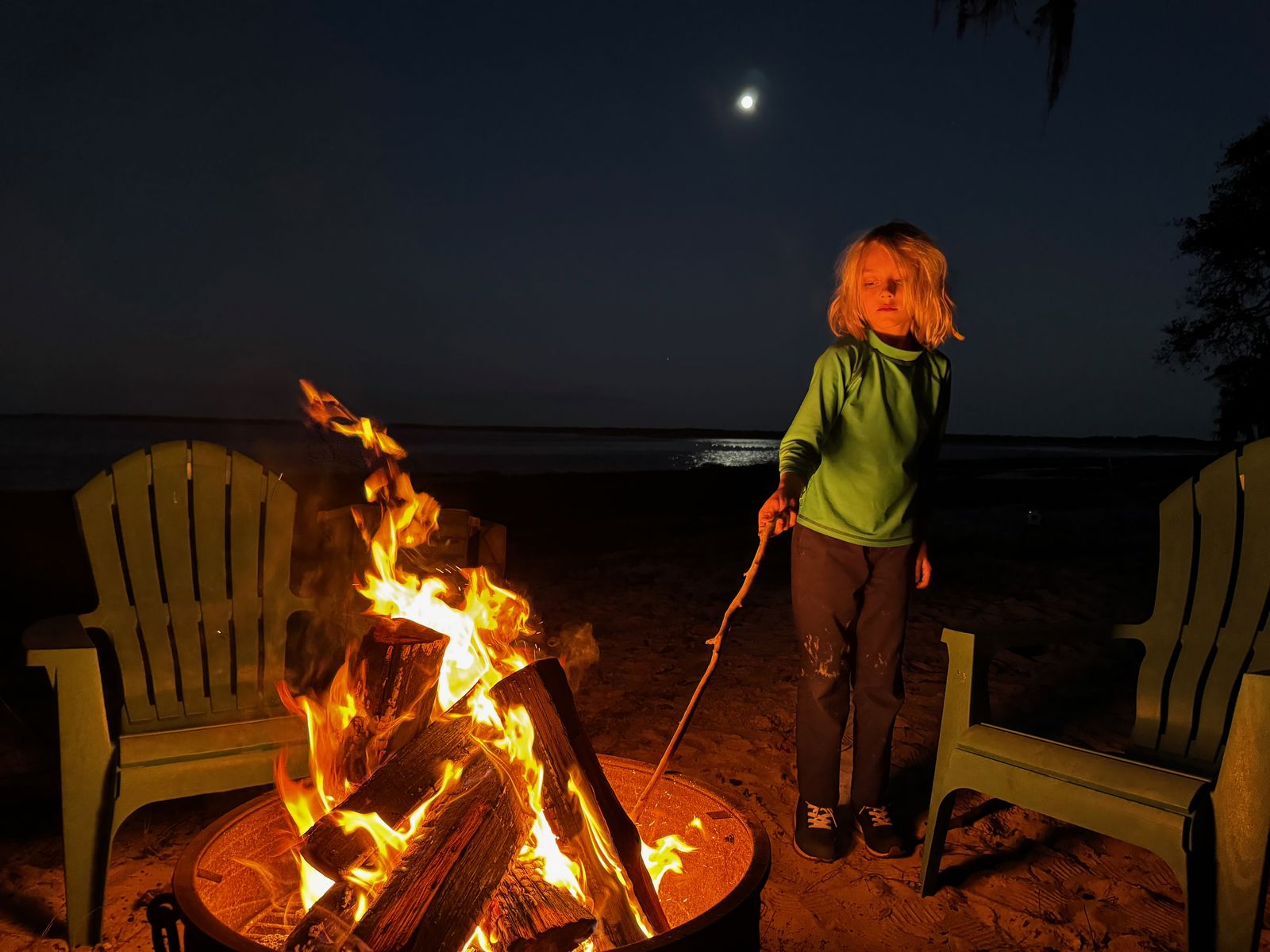 Elijah standing by a bonfire on the shore of the lake behind their house at night, moon reflected on the water